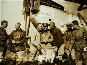 A group of white men conversing in front of a plane named Chicago. The men have their pants tucked into their boots. Some of them are smoking. 